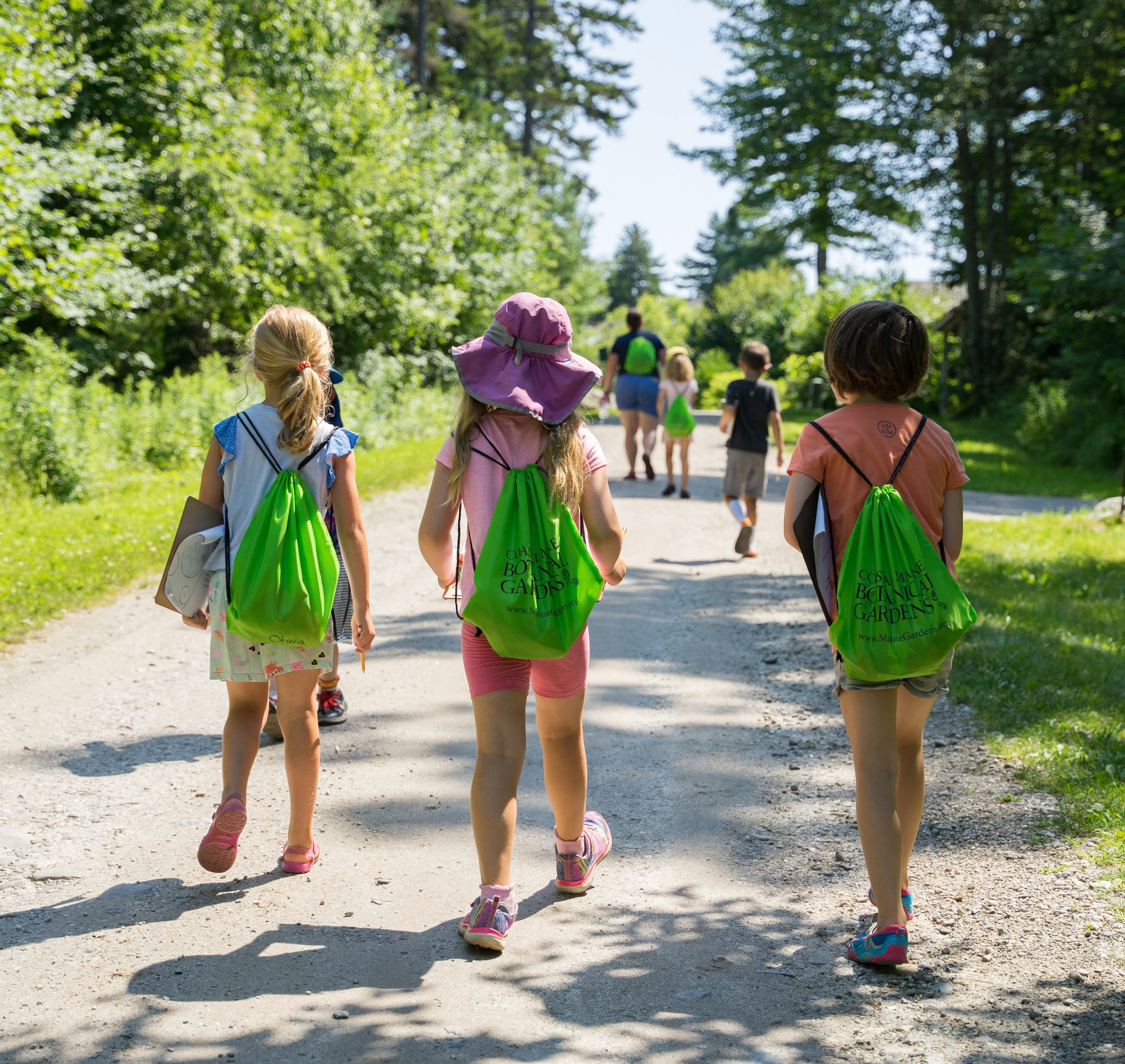 Children walking along a sunny path, each carrying green drawstring backpacks, surrounded by trees and nature.