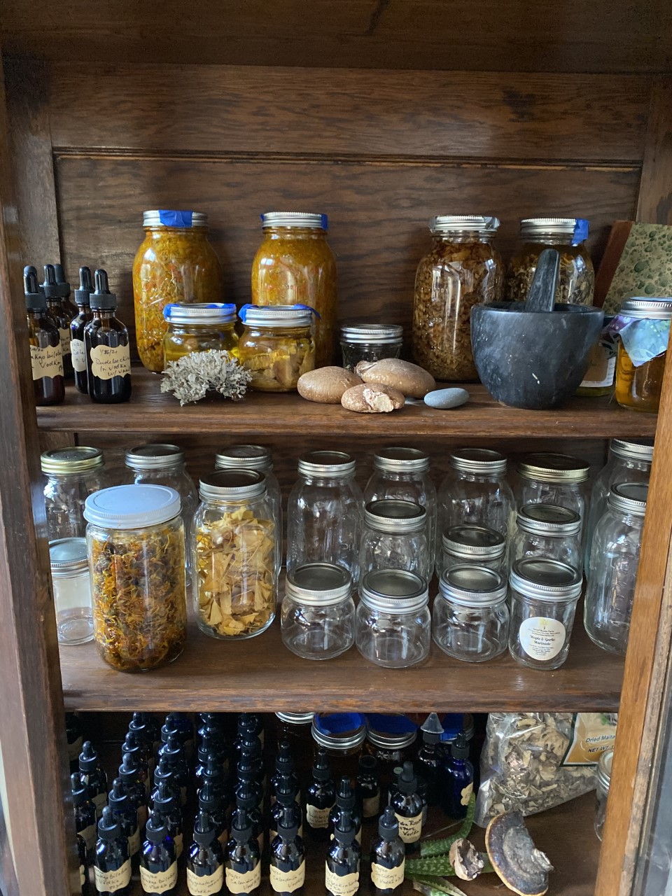A wooden shelf filled with jars of herbs, oils, and dried plants, alongside a mortar and stone.