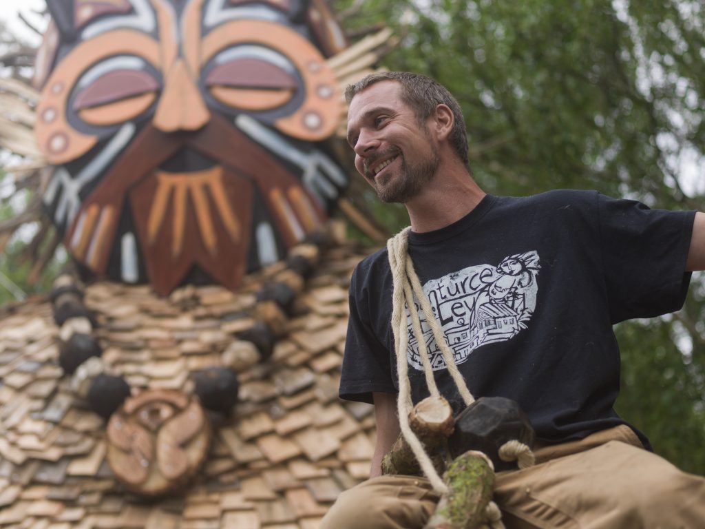 Artist Thomas Dambo smiling while sitting in front of masked giant troll sculpture.