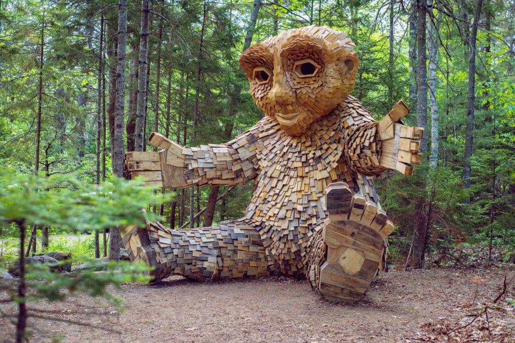A large wooden sculpture of a character with a friendly expression, seated amidst lush green trees in a forest.