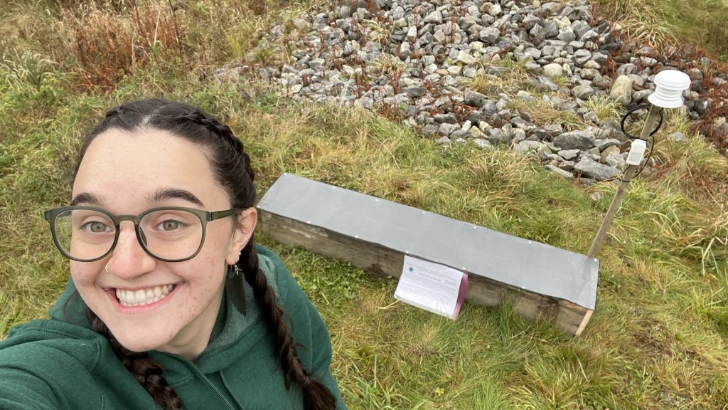 A person with glasses and braided hair smiles at the camera in front of a wooden box with a gray lid and a scientific weather instrument.